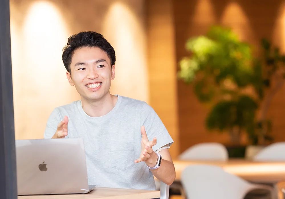 ノートパソコンの前で手を動かしながら笑顔で話す社員。背景は観葉植物のあるカフェスペース